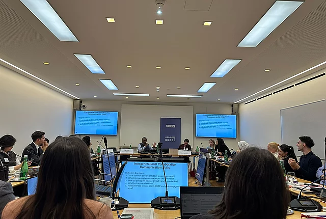 A photo shows the participants of the OSCE young experts event sitting surrounded by screens, during a class on communication skills.