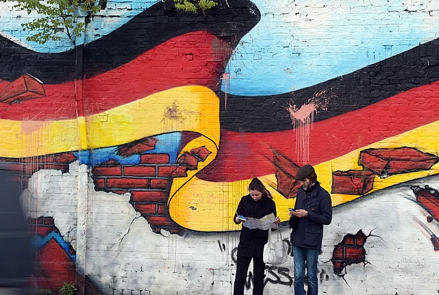 An image shows a photograph of two people, looking into their phones, against the background of a wall with a graffiti of a German flag